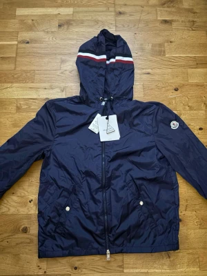 Marinblå vindjacka från Moncler - Snygg marinblå vindjacka från Moncler med huva och dragkedja framtill. Jackan har vita och röda ränder på huvan, två fickor med tryckknappar och Moncler-logga på ärmen. Tillverkad i lätt, glansigt material som står emot vind. Jackan är i storleken M-2. Skriv vid intresse.