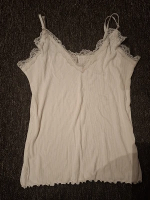 Vit ribbad spetslinne - Säljer ett vitt ribbat linne med smala axelband och v-ringning dekorerad med spets. Linnet har en mjuk och stretchig känsla, perfekt för lager-på-lager eller sommaroutfits. Spetsdetaljerna ger en romantisk touch.