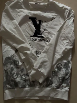 Vit Louis Vuitton sweatshirt - Cool vit sweatshirt från Louis Vuitton med stort svart LV-tryck på bröstet och gråa mönsterdetaljer längst ner. Tröjan har rund hals och långa ärmar, perfekt för dig som gillar exklusiva streetwear-vibes.
