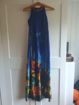 Blå långklänning med blommigt mönster - Säljer en blå långklänning med färgstarkt blommigt mönster nertill. Klänningen har hög halsringning med orange kant och är ärmlös. Perfekt för sommarens alla tillfällen, riktigt flowy och unik design. Stl 36/38. Jag har använt på ett sommarbröllop endast med liten klack, är 163 xm lång. 