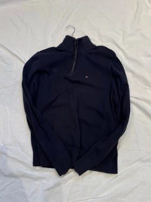 Mörkblå halvzip-tröja Tommy Hilfiger - Mörkblå stickad tröja från Tommy Hilfiger med halvzip och hög krage. Liten broderad logga på bröstet. Tröjan har långa ärmar och ribbade muddar vid ärmslut och nederkant. Perfekt för en stilren och avslappnad look.