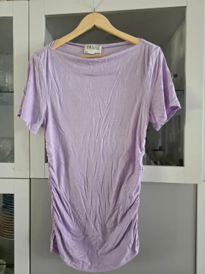 Ljuslila t-shirt från AWARE - Säljer en ljuslila t-shirt från AWARE med korta ärmar och rynkade detaljer på sidan. T-shirten har en avslappnad passform och är tillverkad i mjuk bomull, perfekt för en chill och stilren look.