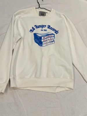 Vit sweatshirt Ed Banger Records - Vit sweatshirt från Ed Banger Records med blå och rosa tryck framtill och text på franska på ryggen. Tröjan har rund hals, långa ärmar och ribbade muddar. Perfekt för dig som gillar streetwear och musikrelaterade plagg.
