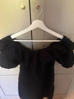 Svart offshoulder kortklänning - Trendig svart kortklänning med offshoulder och puffiga ärmar. Klänningen har en volangdetalj längs axlarna och en figurnära passform. Perfekt för dig som vill ha en stilren och modern look.