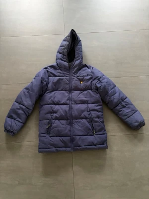 Blå Lyle & Scott pufferjacka med huva - Snygg blå pufferjacka från Lyle & Scott med gul logga på bröstet. Jackan har dragkedja framtill, två fickor med dragkedja (varav en är trasig) och en stor huva. Perfekt för kalla vinterdagar tack vare det vadderade materialet och den långa modellen.