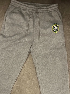 Gråa mjukisbyxor med Brasilien size Xl/L - Säljer ett par gråa mjukisbyxor med Brasilien-logga på vänster lår. Byxorna har dragsko i midjan och sidofickor. Perfekta för chill dagar eller när du vill ha en sportig vibe. Mjukt material som känns skönt mot huden.