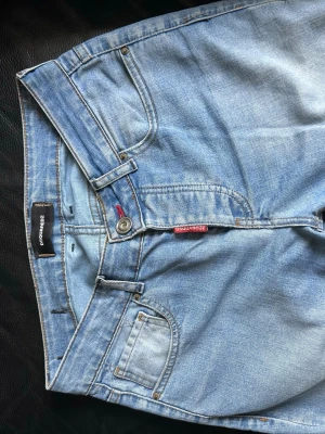 Ljusblå jeans från Dsquared2 - Säljer ett par ljusblå jeans från Dsquared2 med klassisk femficksmodell och röd logotypdetalj vid gylfen. Jeansen har en avslappnad passform och är tillverkade i slitstark denim. Perfekta för en casual och trendig look. Storlekslappen har tyvärr tvättats bort, för omkretsen vid midjan på jeansen eller vid benen så behöver ni dubbla måtten på bilden.