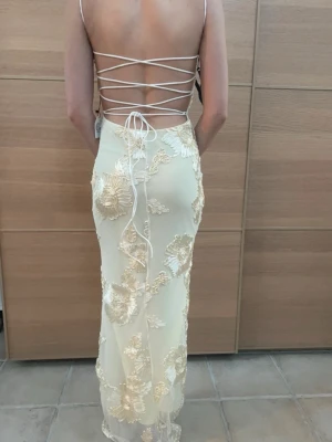 Champagnefärgad långklänning med snörning - Elegant långklänning i champagnefärg med tunna axelband och öppen rygg med snörning. Klänningen har stora blommiga broderier i glansigt material och en figurnära passform. Perfekt för dig som vill sticka ut med en unik och lyxig look.