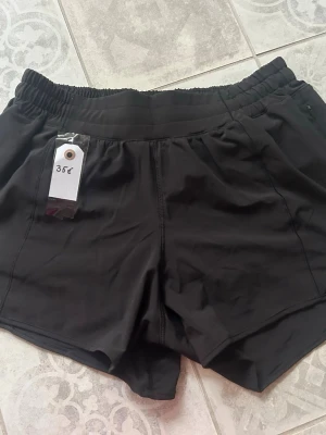 Svarta träningsshorts med resårmidja Lululemon - Snygga svarta träningsshorts från Lululemon med elastisk midja och sportig passform. Shortsen har en enkel design utan synliga loggor och är tillverkade i ett lätt och snabbtorkande material, perfekta för aktiva dagar. Passar dig som vill ha bekväma och stilrena shorts.
