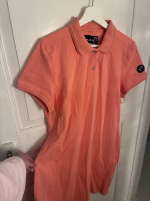 Ljus orange/korall pikéklänning från Pelle P - Säljer en ljus orange/korall pikéklänning från Pelle P med klassisk krage och knappar framtill. Klänningen har korta ärmar och en broderad logga på ärmen. Tillverkad i mjuk bomull som känns skön mot huden. Perfekt för en avslappnad och sportig look.
