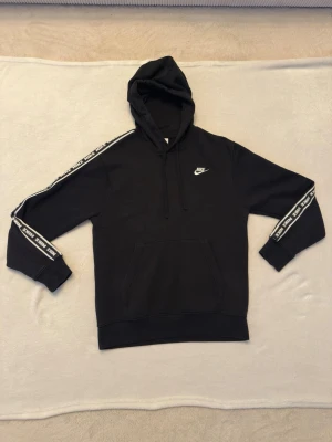 Svart Nike hoodie med logga - Snygg svart hoodie från Nike med vit logga på bröstet och vita Nike-tryck längs ärmarna. Klassisk känguruficka framtill och justerbar huva med snören. Perfekt för en sportig och avslappnad stil.