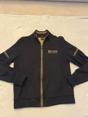 Svart zip hoodie från Hugo Boss - Snygg svart hoodie från Hugo Boss med gulddetaljer och dragkedja framtill. Loggan är broderad på bröstet och insidan av kragen har ett mönster med BOSS-tryck. Ribbade muddar och två fickor framtill. Perfekt för en stilren och sportig look.