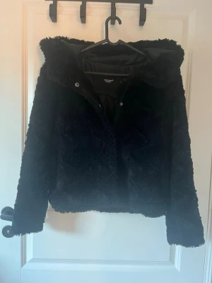 Svart teddyjacka från Vero Moda - Mysig svart teddyjacka från Vero Moda med huva och knäppning framtill. Jackan har fluffig yta och är fodrad på insidan för extra komfort. Perfekt för kyliga dagar när du vill vara både varm och snygg.