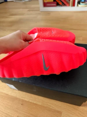 Neonröda Nike tofflor - Säljer ett par neonröda tofflor från Nike med svart Swoosh-logga på sidan och ovansidan. Tofflorna har en unik, räfflad sula och ventilerande hål på ovandelen. Tillverkade i ett mjukt, flexibelt syntetmaterial som känns skönt på foten. Perfekta för chill eller efter träning.