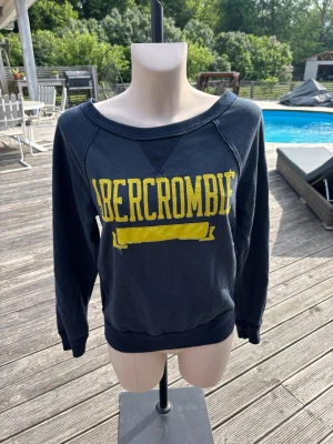 Mörkblå sweatshirt från Abercrombie - Cool mörkblå sweatshirt från Abercrombie med gul text framtill. Tröjan har en avslappnad passform, bred halsringning som kan bäras off-shoulder och långa ärmar med muddar. Perfekt för en chill och sportig look.