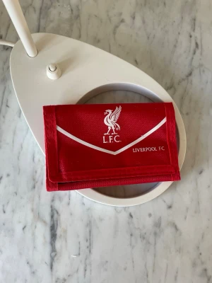 Röd Liverpool FC plånbok - Snygg röd plånbok med Liverpool FC-logga och vit detalj på framsidan. Plånboken har flera fack för kort och sedlar samt stängs med kardborre. Tillverkad i slitstark textil, perfekt för dig som älskar fotboll och vill visa ditt lag.