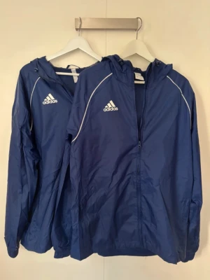 Blå vindjacka från Adidas med huva - Två snygg adidas windbreaker i marinblå och vit färg, dom är i storlek M båda två.Priset är förhandlingsbart så det är bara att buda.