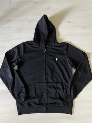 Svart hoodie från Polo Ralph Lauren - Säljer en svart hoodie från Polo Ralph Lauren med dragkedja framtill och klassisk huva. Tröjan har det ikoniska vita Polo-logotrycket på bröstet och två fickor framtill. Perfekt för en chill och stilren look. Storlek S/M står M känns som S