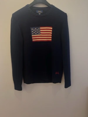 Mörkblå stickad tröja Polo Ralph Lauren - S (barn XL) Mörkblå stickad tröja från Polo Ralph Lauren med amerikansk flagga i rött, vitt och blått på bröstet. RL-broderi i rött vid nederkanten. Klassisk rund halsringning och långa ärmar. Perfekt för dig som gillar preppy stil.