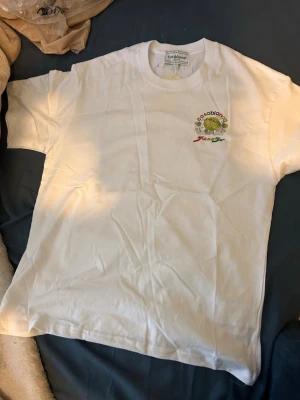 Vit Casablanca t-shirt med tryck - Säljer en vit t-shirt från Casablanca med färgglatt tryck på bröstet. T-shirten har rund hals och korta ärmar. Materialet är mjuk bomull och passformen är normal. Perfekt för dig som gillar stilrena plagg med en twist.