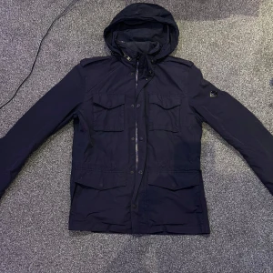 Marinblå jacka från J.Lindeberg - Snygg marinblå field jacket från J.Lindeberg med huva och flera praktiska fickor. Jackan har en stilren design med dragkedja och knappar framtill. Perfekt för höst och vår men även sommar.