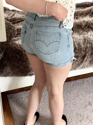 Ljusblå denimshorts med nitar - Säljer ett par ljusblå denimshorts från Levi's. Shortsen har slitna detaljer, råa kanter och silverfärgade nitar på bakfickorna och sidorna. Perfekta för sommaren och ger en cool, avslappnad vibe.