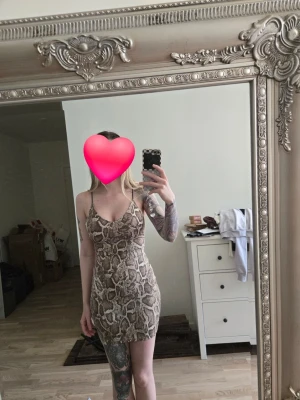 Beige kort klänning med ormmönster - Säljer en beige kort klänning med ormmönster och smala axelband. Klänningen har en tight passform och är ärmlös, perfekt för dig som vill sticka ut. Materialet känns mjukt och stretchigt, troligen polyester eller syntet. Helt nytt med prislapp.