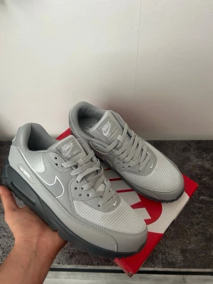 Nike Air Max 90 grå sneakers - Nike Air Max 90 sneakers i helgrå färg med vit swoosh och detaljer. Ovandelen är i mesh och syntet, med klassisk Air Max-dämpning i sulan. Skorna har en chunky siluett och tydliga paneler, samt snörning framtill. Perfekta för dig som gillar stilrena och sportiga sneakers.