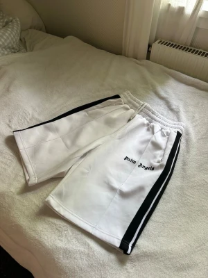 Vita shorts från Palm Angels - Palm Angels vita shorts med svarta sidoränder och broderad logga på vänster ben. De har elastisk midja och sportig look, perfekta för en avslappnad stil. Materialet känns mjukt och lätt, troligen polyester.