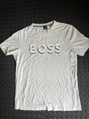 Vit BOSS t-shirt med stor logga - Säljer en vit t-shirt från BOSS med stor, tydlig logga i svart och vitt på bröstet. Klassisk rund halsringning och korta ärmar. Tillverkad i mjuk bomull som känns skön mot huden. Perfekt för dig som gillar stilrena och enkla plagg med tydlig branding.