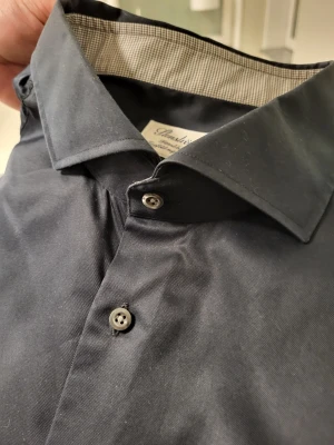 Stenströms fitted body two fold super svart skjorta, storlek 46 18 - Klassisk svart skjorta från Stenströms med lång ärm och knappar framtill. Skjortan har en stilren krage och är tillverkad i ett mjukt bomullstyg. Perfekt för en clean och snygg look.