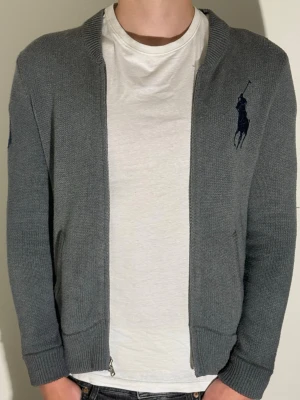 Grå ziptröja från Ralph Lauren - Snygg grå ziptröja från Ralph Lauren med mörkblå logga broderad på bröstet. Tröjan har ribbade muddar, rak passform och dragkedja framtill. Perfekt att slänga på sig över en t-shirt för en clean och stilren look. Finns tyvärr ingen storleks lapp men killen på bilden har vanligtvis M så skulle tro att tröja passar best för dom som har S