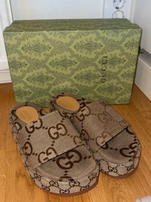 Gucci beige platåtofflor med GG-mönster - Säljer ett par trendiga beige platåtofflor från Gucci med det klassiska GG-monogrammet i brunt. Tofflorna har bred rem över foten och är tillverkade i canvas med läderdetaljer. Perfekta för dig som vill ha en statement-look och älskar lyxiga accessoarer, flera storlekar finns inne, och pris kan diskuteras vid snabb affär 🌸