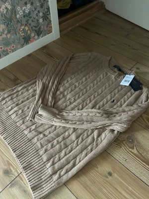 Beige kabelstickad tröja Polo Ralph Lauren - Snygg beige kabelstickad tröja från Polo Ralph Lauren med rund halsringning och klassisk broderad logga på bröstet. Tröjan har ribbade muddar vid ärmslut och nederkant. Perfekt för lager på lager och en stilren look. Säljes billigt pga fel storlek för mig ocy tunt material passar L