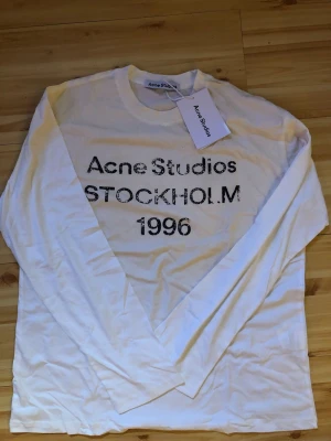 Vit långärmad t-shirt Acne Studios - Vit långärmad t-shirt från Acne Studios med svart tryck på bröstet där det står 'Acne Studios STOCKHOLM 1996'. Klassisk rund halsringning och mjukt material, perfekt för en clean och stilren look.