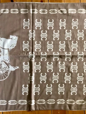 Beige sjal från Celine med logomönster - Snygg beige sjal från Celine med ikoniskt logomönster i vitt och dekorativt häst- och vagnmotiv. Sjalen har en klassisk rektangulär form och är tillverkad i ett mjukt material som känns lyxigt. Perfekt accessoar för att lyfta din outfit. Kashmir.