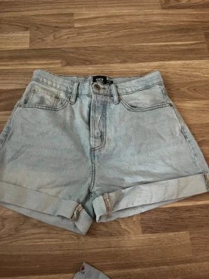 Ljusblå denimshorts från 157 - Säljer ett par ljusblå denimshorts från 157 med hög midja och uppvikta benslut. Klassisk femficksmodell med bälteshällor och knappgylf. Perfekta till sommaren för en avslappnad och trendig look.