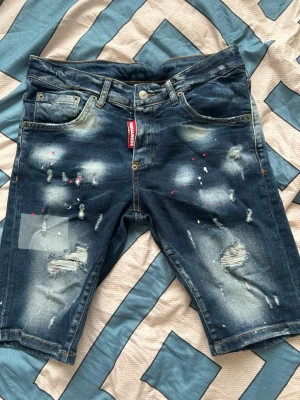 Blå distressed jeansshorts ICON - Säljer ett par blå jeansshorts från ICON med slitningar, patchdetaljer och färgstänk framtill. Klassisk femficksmodell med knappgylf och bälteshällor. Bakfickorna är markerade och det finns en stor orange ICON-lapp bak i midjan. Materialet är denim.