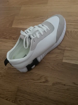Vita sneakers med grå detaljer - Snygga vita sneakers från Axel Arigato med grå mockadetaljer och svarta detaljer vid hälen. Skon har en chunky, räfflad sula och klassisk snörning. Perfekt för dig som gillar stilrena och moderna sneakers.