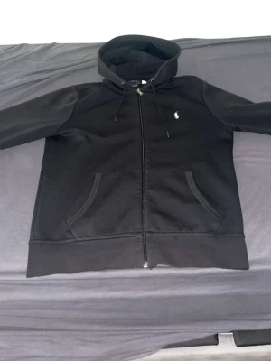 Svart hoodie från Ralph Lauren - Säljer en svart hoodie från Ralph Lauren med dragkedja framtill och två fickor. Klassisk vit logga på bröstet och justerbar huva med snören. Tillverkad i mjukt bomullsmaterial som känns skönt mot huden. Perfekt för en avslappnad och stilren look.