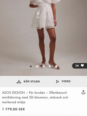 Elfenbensvit miniklänning ASOS DESIGN - Elfenbensvit kortklänning från ASOS DESIGN med markerad midja, stråveck och 3D-blommor. Klänningen har v-ringning, volangdetaljer och puffiga korta ärmar i transparent material. Perfekt för dig som vill ha en romantisk och unik look. Helt oanvänd då jag beställde två i olika storlekar och kommer behålla den andra.