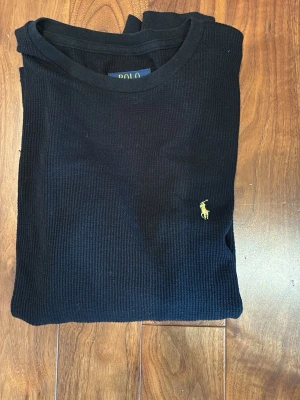 Svart långärmad tröja Polo Ralph Lauren - Klassisk svart långärmad tröja från Polo Ralph Lauren med rund hals och diskret gult broderat logotyp på bröstet. Tröjan är ribbstickad och har en smal passform som passar perfekt till jeans eller chinos. Storlek S!