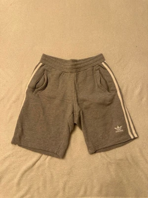 Grå Adidas shorts med vita ränder - Gråa shorts från Adidas med klassiska vita ränder längs sidorna och broderad logga framtill. Mjukt material med två fickor fram och elastisk midja för skön passform. Perfekta för en avslappnad stil.