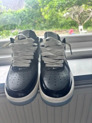 Nike Air Force 1 svart/vit sneakers - Snygga Nike Air Force 1 sneakers i svart och vitt med klassisk låg siluett. Skorna har vita tjocka skosnören, perforerad tåbox och vit sula med blåaktig yttersula. Tillverkade i läder med textildetaljer och ikonisk Nike Air-logga på plösen.