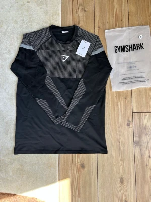 Svart långärmad träningströja Gymshark - Snygg svart långärmad träningströja från Gymshark med gråmelerade detaljer på axlar, bröst och ärmar. Tillverkad i ett stretchigt funktionsmaterial som andas, perfekt för gymmet eller löpning. Gymshark-logga på bröstet och modern passform. Storlek är M och passar mellan 170-184 skulle jag säga, lite liten för mig är 190 o väger 86