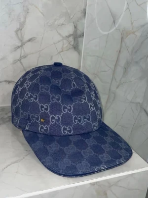 Blå GG-monogram keps från Gucci - Snygg blå keps från Gucci med klassiskt GG-monogram i ljusgrått över hela kepsen. Kepsen har böjd skärm och är tillverkad i denimliknande material. Perfekt accessoar för att lyfta din streetwear-look.