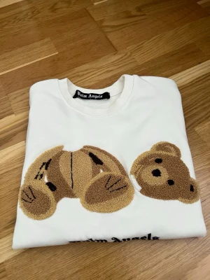 Vit sweatshirt med nalle från Palm Angels - Säljer en vit sweatshirt från Palm Angels med ett stort broderat motiv av en nallebjörn i beige och brunt på framsidan. Tröjan har rund halsringning och svart Palm Angels-logga under motivet. Perfekt för dig som gillar streetwear och unika detaljer.