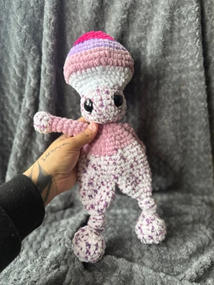 Handvirkad rosa svampfigur - Unik handvirkad mjukis i form av en svampfigur med stora svarta ögon. Färgerna går i rosa, vitt, lila och rött. Tillverkad i mjukt garn med fluffig textur och detaljerade ben och armar. Perfekt som dekoration eller gosedjur.