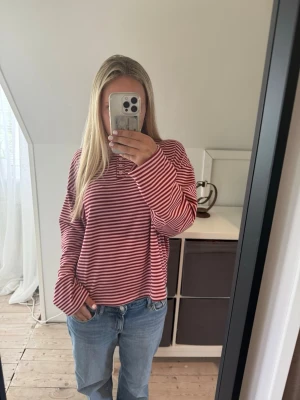 Randig röd långärmad tröja - Säljer en röd och vit randig långärmad tröja med rund hals och knappar vid halsringningen. Tröjan har en avslappnad passform och är perfekt för en chill vardagslook. Materialet känns mjukt och bekvämt mot huden. Tröjan har en stark doft ( inget som luktar äckligt men som en stark parfym), vilket den gjorde redan när jag köpte den. 