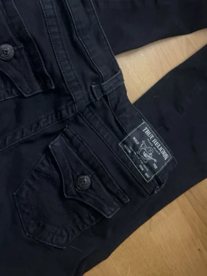 Svarta True Religion low rise jeans - Säljer ett par svarta True Religion Jeans i Modell JOEY Low Rise Flare i storlek 23. Dom har slitningar längst ner vid fötterna men annars i bra skick.